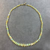 A beaded necklace made of small, irregular opal-like beads showing iridescent colors of yellow, green, and white, with a gold clasp, displayed on a textured gray background.