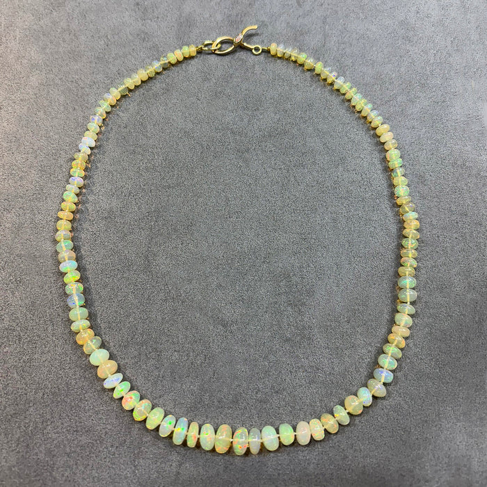 A beaded necklace made of small, irregular opal-like beads showing iridescent colors of yellow, green, and white, with a gold clasp, displayed on a textured gray background.